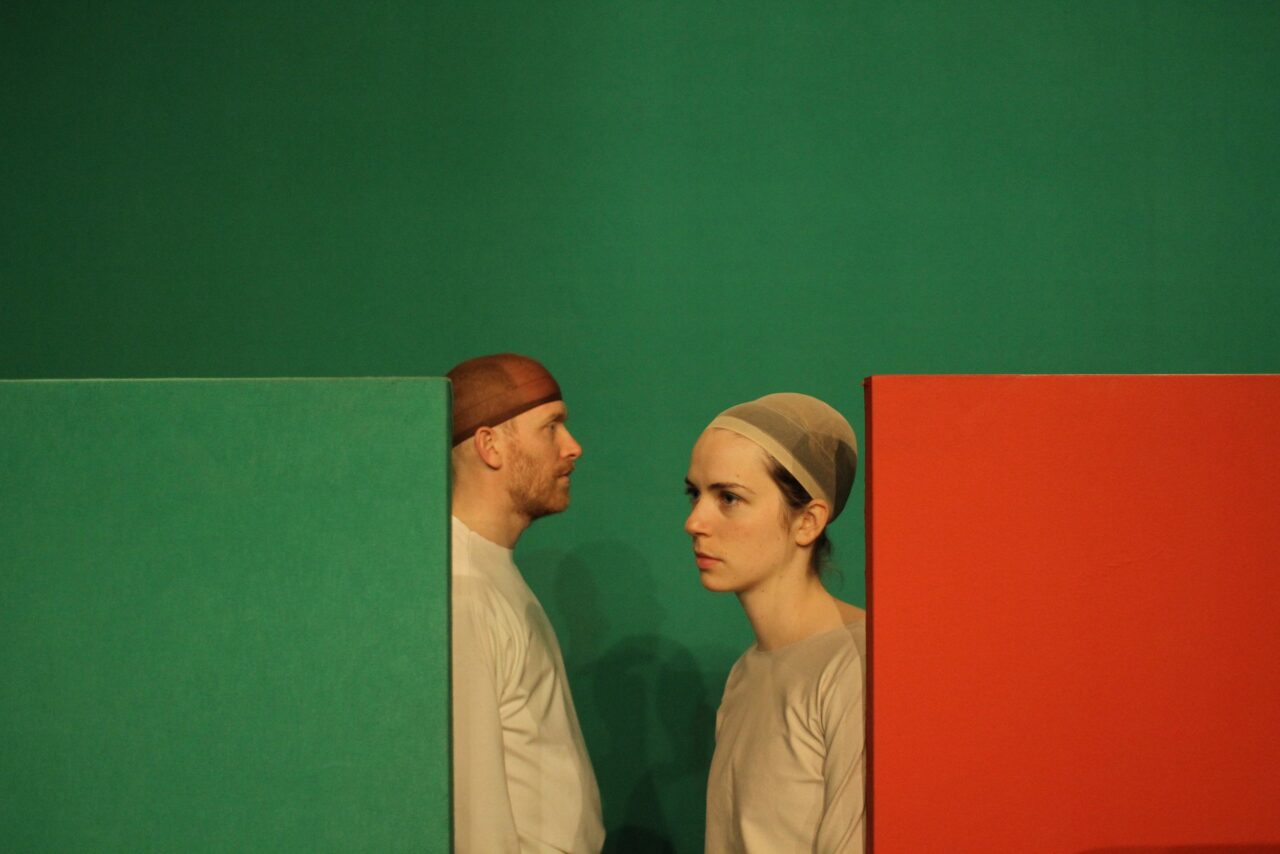 Zwei Personen mit beigefarbenen Kostümen und eng anliegenden Hauben stehen zwischen farbigen Wänden. Links ein Mann vor einer grünen Wand, rechts eine Frau vor einer roten Wand.