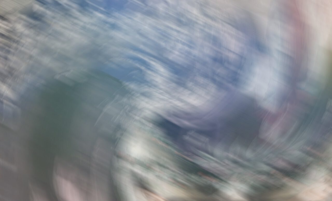 Das Bild zeigt eine abstrakte, verschwommene Darstellung in Blau-, Grau-, Grün- und Beigetönen. Die Farben und Formen verlaufen ineinander. Durch die Bewegungsunschärfe entsteht ein dynamischer, beinahe wirbelnder Eindruck. Die Darstellung wirkt weich und atmosphärisch. Konkrete Gegenstände oder Personen sind nicht eindeutig erkennbar.