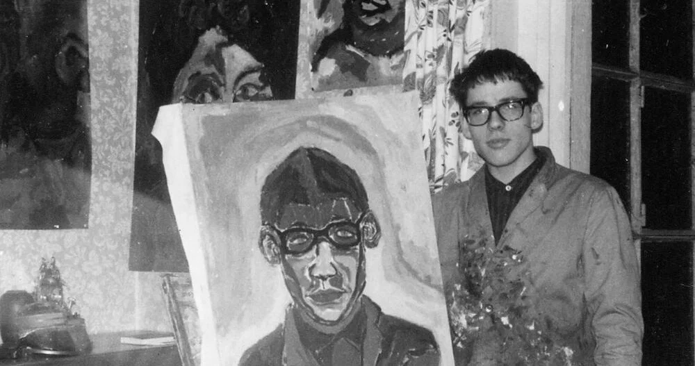 Schwarz-Weiß-Fotografie eines jungen Mannes mit Brille, der neben einem großen Porträtgemälde steht. Der Mann trägt ein dunkles Hemd und eine helle Jacke und hält einen Blumenstrauß vor sich. Auf dem Gemälde ist ein Mann mit dunklem Haar und auffälliger schwarzer Brille frontal dargestellt. Im Hintergrund sind mehrere weitere Porträts und Gemälde zu sehen, die teilweise an einer Wand lehnen. Die Aufnahme wirkt wie eine historische Fotografie aus einer privaten Atelier- oder Ausstellungssituation.