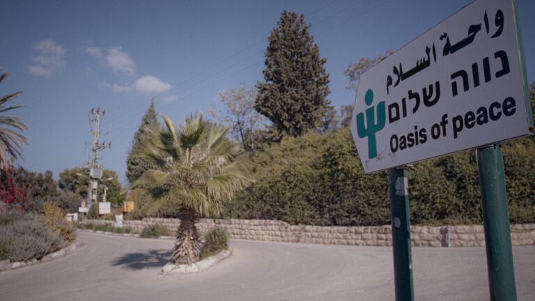 Ein Straßenschild am Rand einer ruhigen Straße weist auf den Ort „Oasis of Peace“ hin. Auf dem Schild stehen der Name auf Englisch, Hebräisch und Arabisch sowie ein grünes Symbol mit stilisierten Figuren. Die Straße verläuft leicht gebogen durch eine grüne Umgebung mit Palmen, Bäumen und Sträuchern. Links ist ein kleiner Kreisverkehr mit einer Palme in der Mitte zu sehen. Im Hintergrund stehen weitere Bäume sowie ein Strommast. Der Himmel ist blau mit wenigen Wolken, und die Szene wirkt ruhig und sonnig.