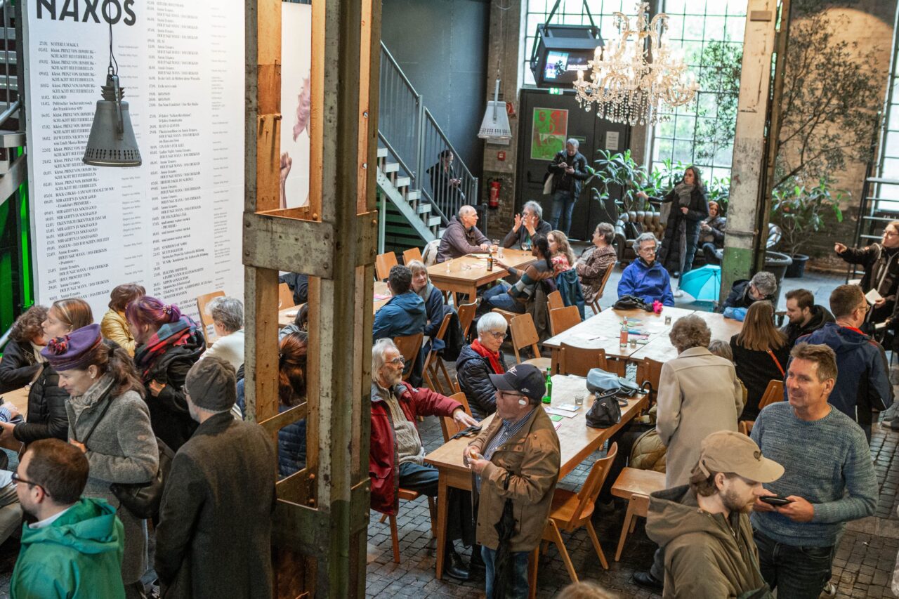 Menschen sitzen und stehen in einem Foyer mit langen Holztischen, im Hintergrund hängen ein Kronleuchter und große Pflanzen.