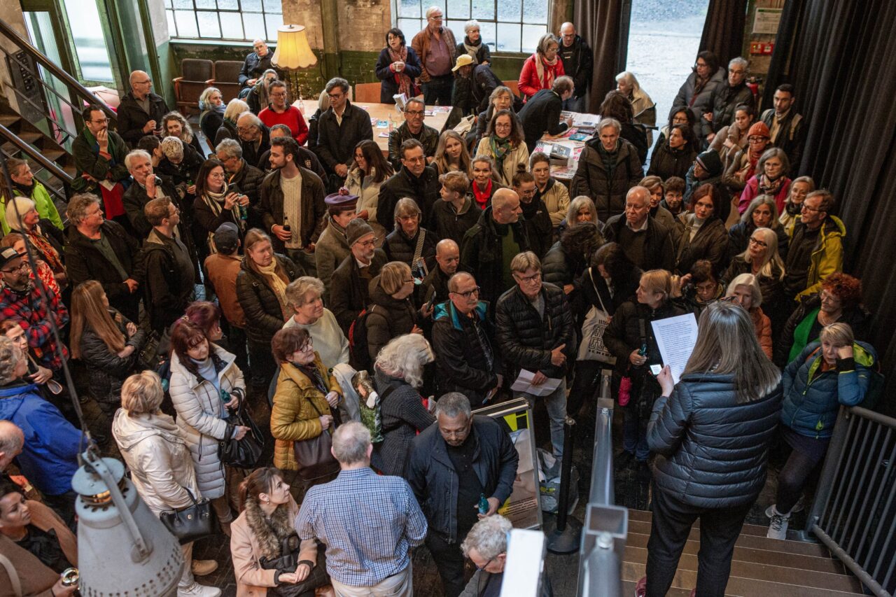 Große Menschenmenge in einem Foyer, eine Frau liest von einem Blatt, während die Gruppe zuhört.