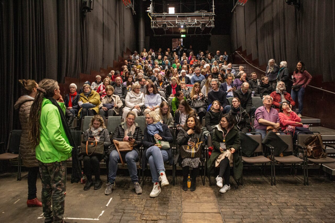 Großes Publikum sitzt in einem Theatersaal, einige Menschen im Vordergrund unterhalten sich, andere schauen auf ihre Handys.