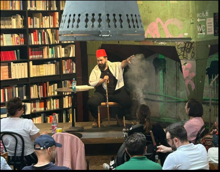 Ein Erzähler mit rotem Fez-Hut sitzt auf einer kleinen Bühne inmitten eines Cafés mit Bücherregalen und spricht vor Publikum.