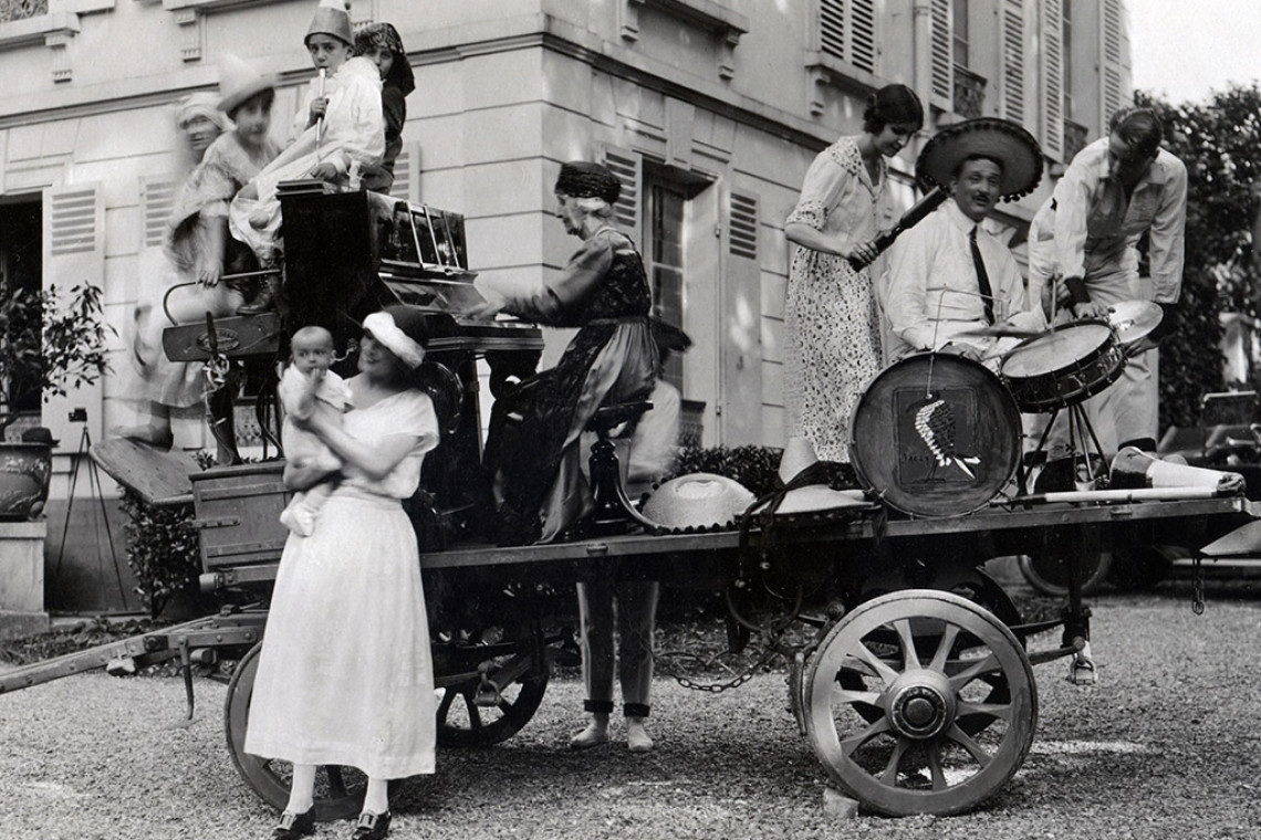 Schwarz-Weiß-Foto: Menschen musizieren und feiern auf einem Wagen vor einem Haus; eine Frau spielt Klavier, ein Mann Schlagzeug, andere tanzen, eine Frau hält ein Baby im Arm.