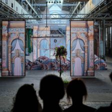 In einer großen Industriehalle (der Naxoshalle) steht eine kunstvolle Bühneninstallation mit drei hohen, bemalten Kulissen, die wie dekorative Türen oder Fenster gestaltet sind. In der Mitte hängt ein großer, funkelnder Kronleuchter über stilisierten Wellenformen und Blumen. Im Vordergrund sind die dunklen Silhouetten von Zuschauern zu sehen.