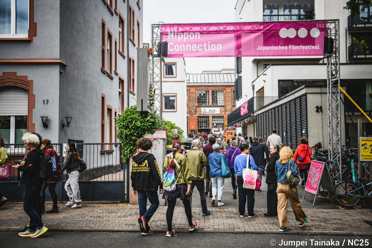 Straßenszene vor dem Veranstaltungsort. Ein großes Banner mit der Aufschrift „Nippon Connection – Japanisches Filmfestival“ spannt sich über die Straße. Zahlreiche Besucher*innen gehen darunter hindurch Richtung Gelände, umgeben vonWohn- und Industriegebäuden.