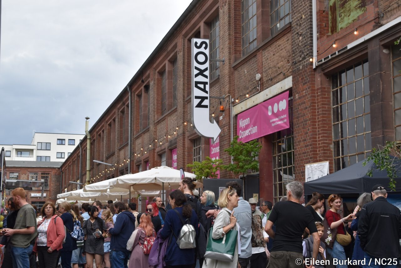 Vor einem langen Backsteingebäude mit großen Fenstern hängt ein vertikales Schild mit der Aufschrift „NAXOS“. Davor drängt sich eine größere Menschenmenge auf einer Straße oder einem Hof. Viele Besucher*innen stehen in Gruppen zusammen, unterhalten sich oder warten. Weiße Sonnenschirme und Stände deuten auf ein Festival hin. An der Fassade hängen pinke Banner mit der Aufschrift „Nippon Connection“. Die Stimmung wirkt lebendig und gesellig.