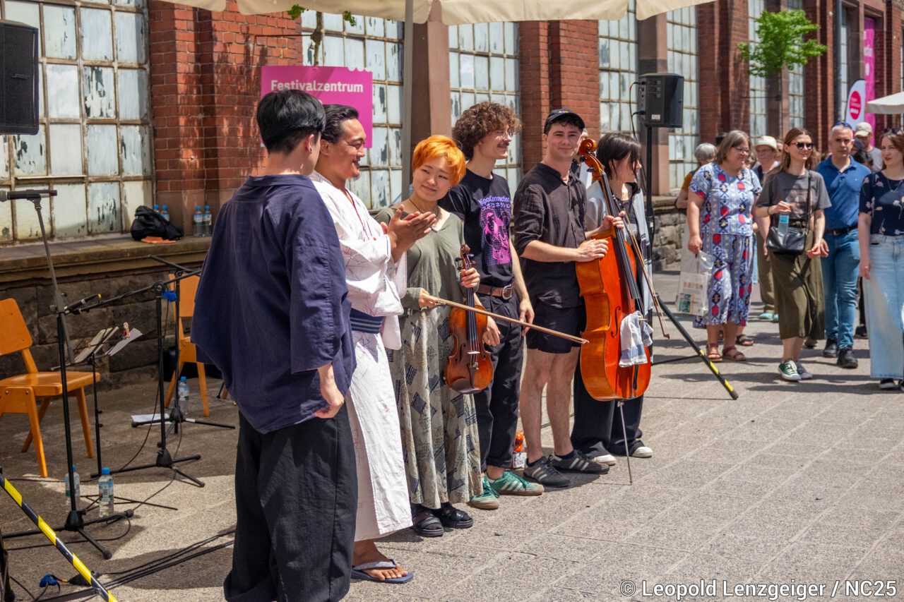 Mehrere Musiker*innen stehen nebeneinander vor einem Backsteingebäude und schauen in Richtung Publikum. Einige halten Instrumente wie Violine und Cello in der Hand. Eine Person klatscht, andere lächeln. Im Hintergrund stehen weitere Besucher*innen, einige beobachten die Szene. Mikrofone, Stühle und Lautsprecher sind aufgebaut. Ein pinkes Schild mit der Aufschrift „Festivalzentrum“ ist an der Wand zu sehen. Die Szene vermittelt den Abschluss oder Applaus nach einem Auftritt im Freien.