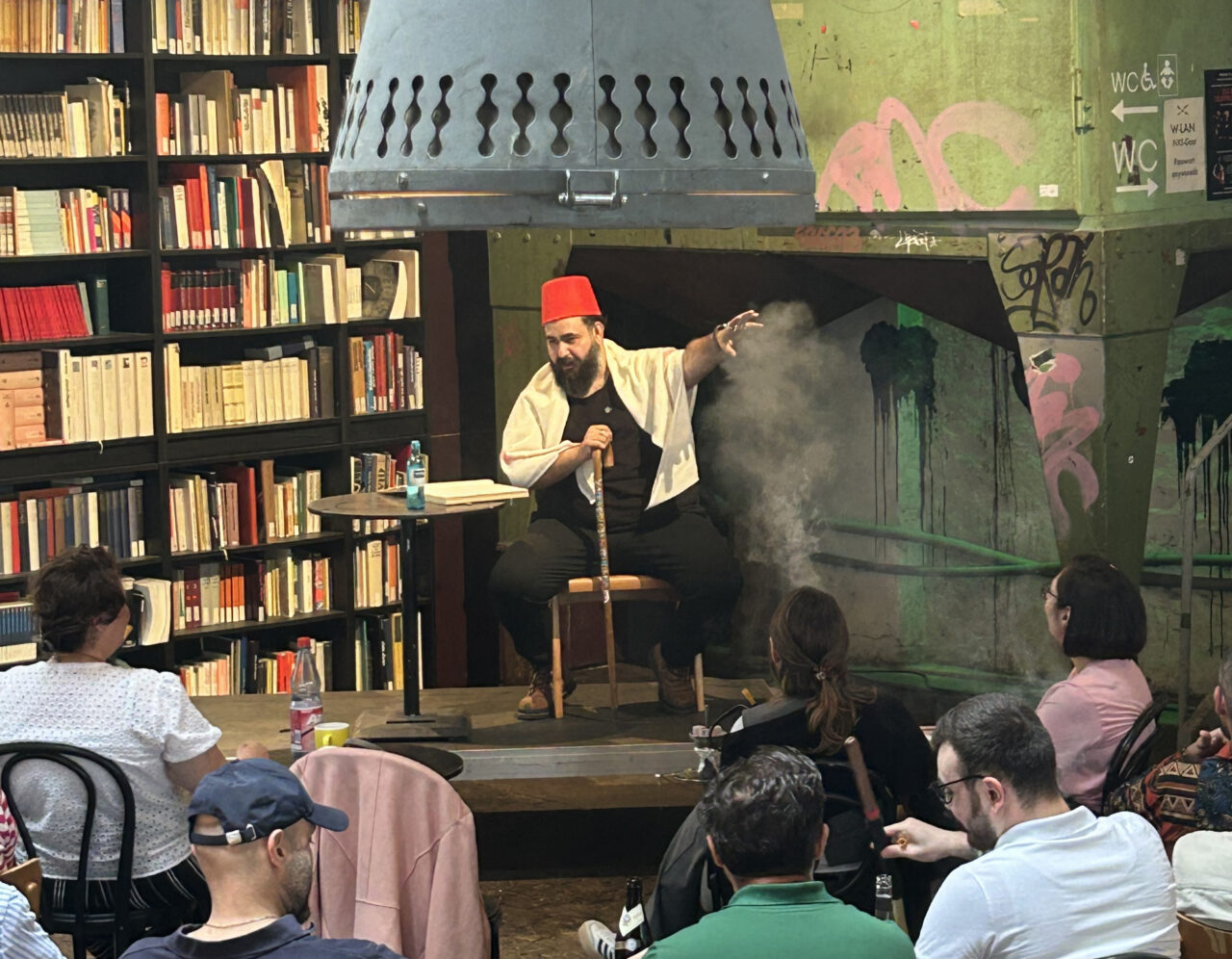 A storyteller wearing a red fez sits on a small stage in a café with bookshelves, speaking to an audience.