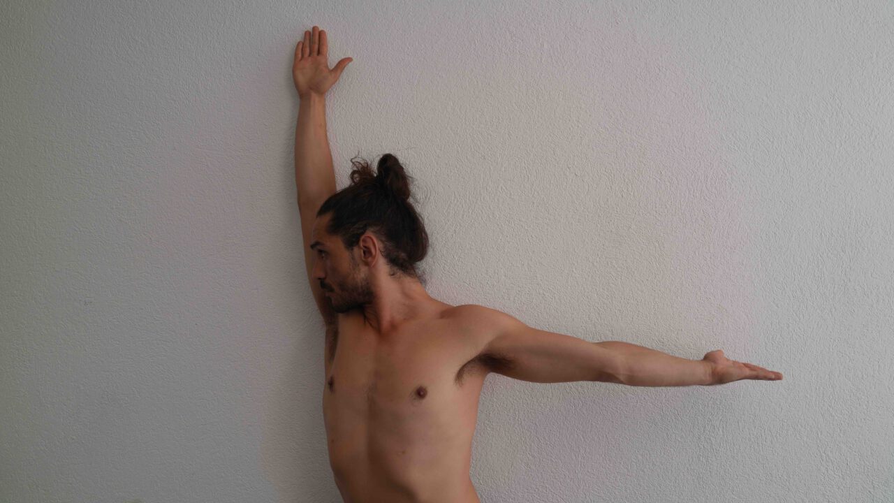 Ein shirtloser Mann mit dunklem Haar, das zu einem Dutt gebunden ist, steht seitlich vor einer hellen Wand. Ein Arm ist senkrecht nach oben ausgestreckt und die Hand berührt die Wand. Der andere Arm ist waagerecht zur Seite ausgestreckt. Sein Kopf ist zur Seite gedreht und sein Blick geht nach unten.