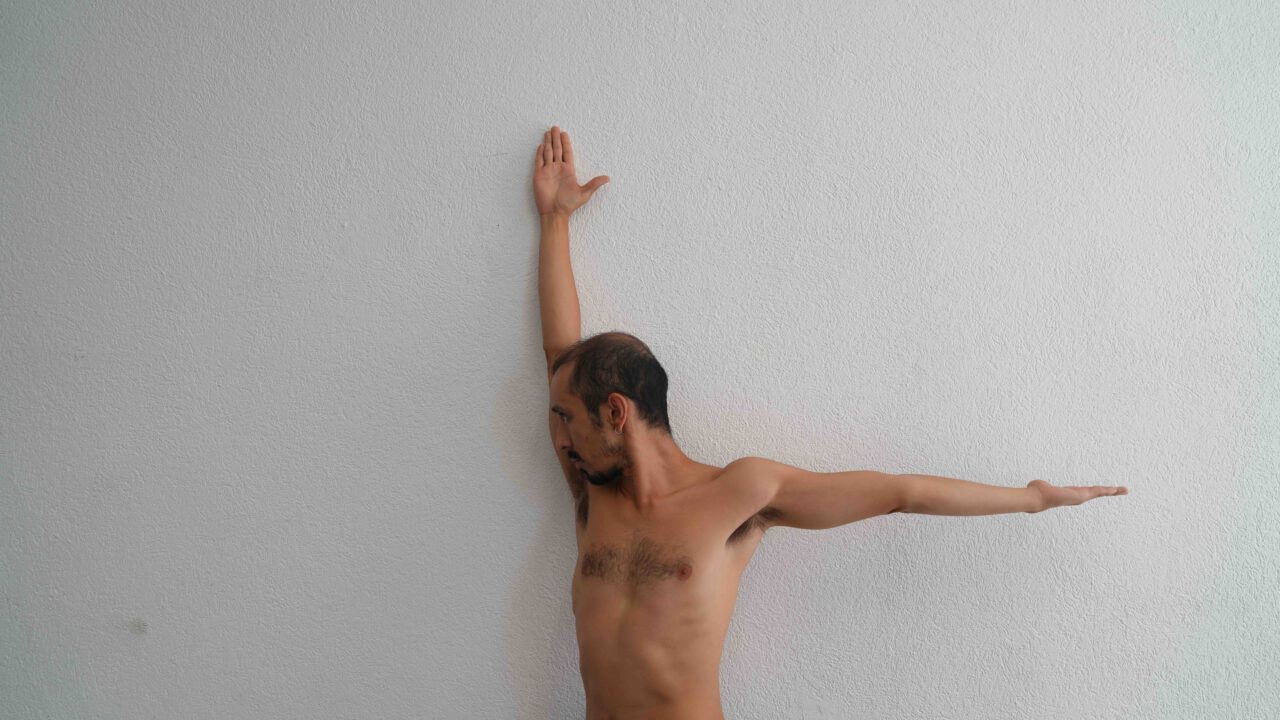 Ein shirtloser Mann steht seitlich vor einer hellen Wand. Ein Arm ist senkrecht nach oben ausgestreckt und die Hand berührt die Wand. Der andere Arm ist waagerecht zur Seite ausgestreckt. Sein Kopf ist zur Seite gedreht und sein Blick geht nach unten. Er trägt einen kurzen Bart und hat dunkles, kurzes Haar.