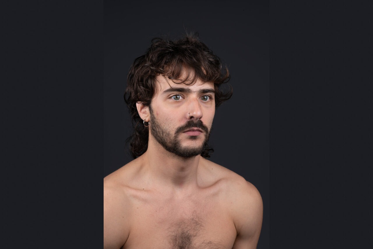 Portrait of a young man against a black background. He has dark, curly hair, a short beard, and piercings on his nose and ear. He looks directly forward with a calm and attentive expression. His shoulders and upper torso are visible.