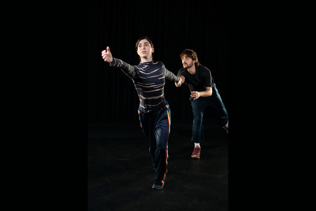 Two dancers move across a dark stage. In the foreground, one person stretches an arm far forward while the second person behind them mirrors a similar movement. The stage lighting highlights the dynamic gestures and creates a focused, artistic atmosphere.