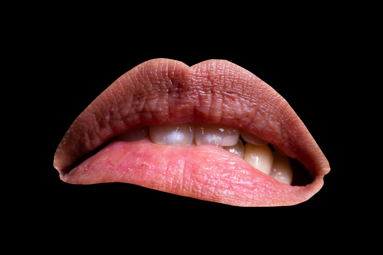 Nahaufnahme eines leicht geöffneten Mundes vor schwarzem Hintergrund. Die Lippen sind rosafarben und deutlich sichtbar strukturiert. Die Zunge berührt die oberen Schneidezähne. Ein Teil der weißen Zähne ist sichtbar. Das Bild wirkt wie eine Studioaufnahme mit starkem Kontrast zwischen Hauttönen und schwarzem Hintergrund.
