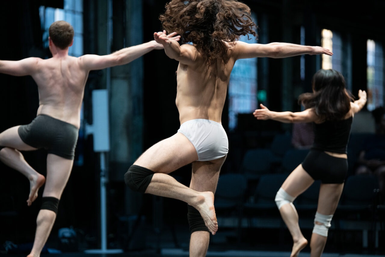 Drei Tänzerinnen und Tänzer sind barfuß während einer Performance in einer ehemaligen Industriehalle zu sehen. Die Aufnahme zeigt sie von hinten in einer synchron wirkenden Bewegung mit ausgestreckten Armen und angehobenen Beinen. Schlichte Kostüme und die industrielle Architektur lenken den Blick auf die Körper, die Bewegung und die räumliche Wirkung der Choreografie.
