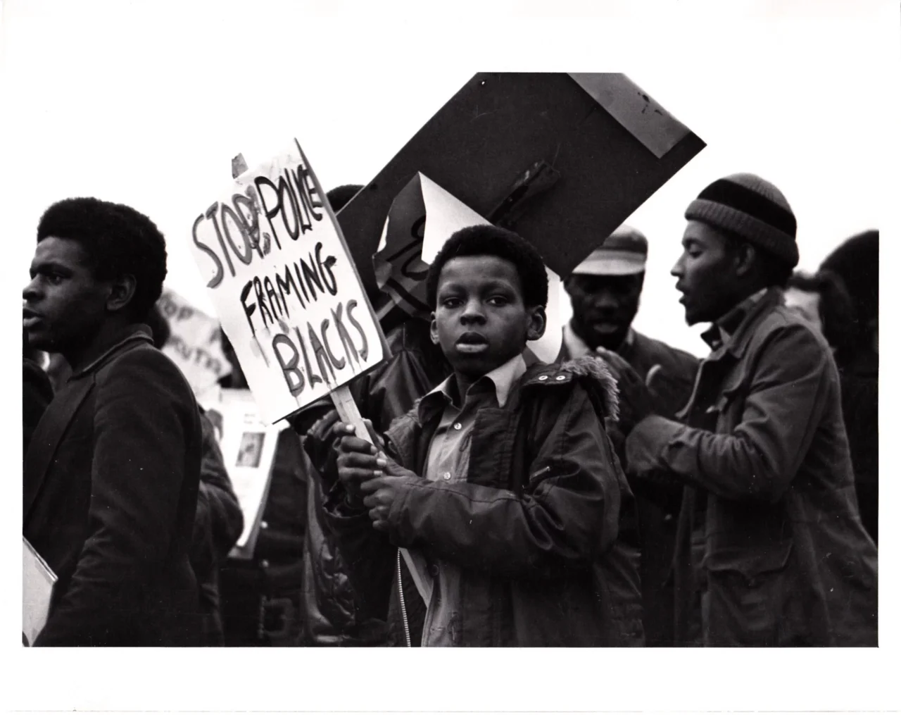Das Schwarz-Weiß-Foto zeigt eine Gruppe von Menschen bei einer Demonstration. Im Mittelpunkt steht ein junger Schwarzer Junge in einer Jacke. Er hält mit beiden Händen ein Protestschild, auf dem „Stop Police Framing Blacks“ geschrieben steht. Sein Blick ist auf die Kamera gerichtet. Links und rechts von ihm stehen weitere Demonstrierende, teilweise im Gespräch, teilweise mit weiteren Schildern.
