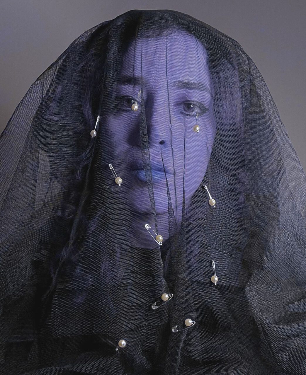 Ein Gesicht hinter einem schwarzen Schleier, in dem mehrere Sicherheitsnadeln mit Perlen befestigt sind; der Ausdruck wirkt ernst und melancholisch.