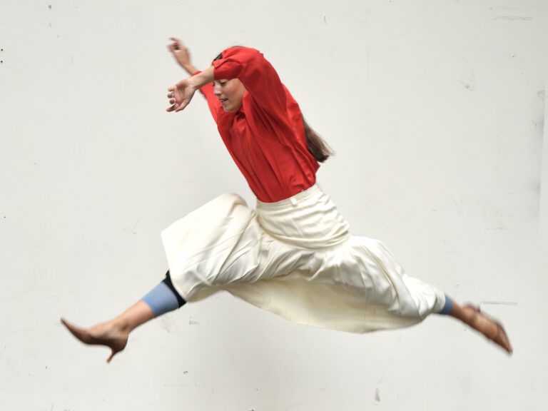A dancer leaps dynamically through the air, wearing a red top and a flowing white skirt.