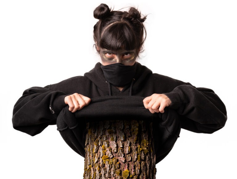 Das Bild zeigt eine Person vor einem neutralen, hellgrauen Hintergrund. Sie trägt einen schwarzen Hoodie und hat den unteren Teil des Gesichts mit einem schwarzen Tuch oder einer Maske bedeckt. Die dunklen Haare sind zu zwei kleinen Knoten auf dem Kopf frisiert, mit geradem Pony vorne. Auffällig ist das Make-up: Um die Augen herum ist dunkle Farbe aufgetragen, ergänzt durch goldene Akzente, was dem Blick eine intensive, fast geheimnisvolle Wirkung verleiht. Die Person schaut direkt in die Kamera. Mit beiden Händen hält sie den schwarzen Hoodie, der über einen Baumstamm gespannt ist. Der Baumstamm mit seiner rauen, strukturierten Rinde bildet einen starken Kontrast zur Kleidung und zum glatten Hintergrund. Insgesamt wirkt die Inszenierung künstlerisch und leicht mystisch.