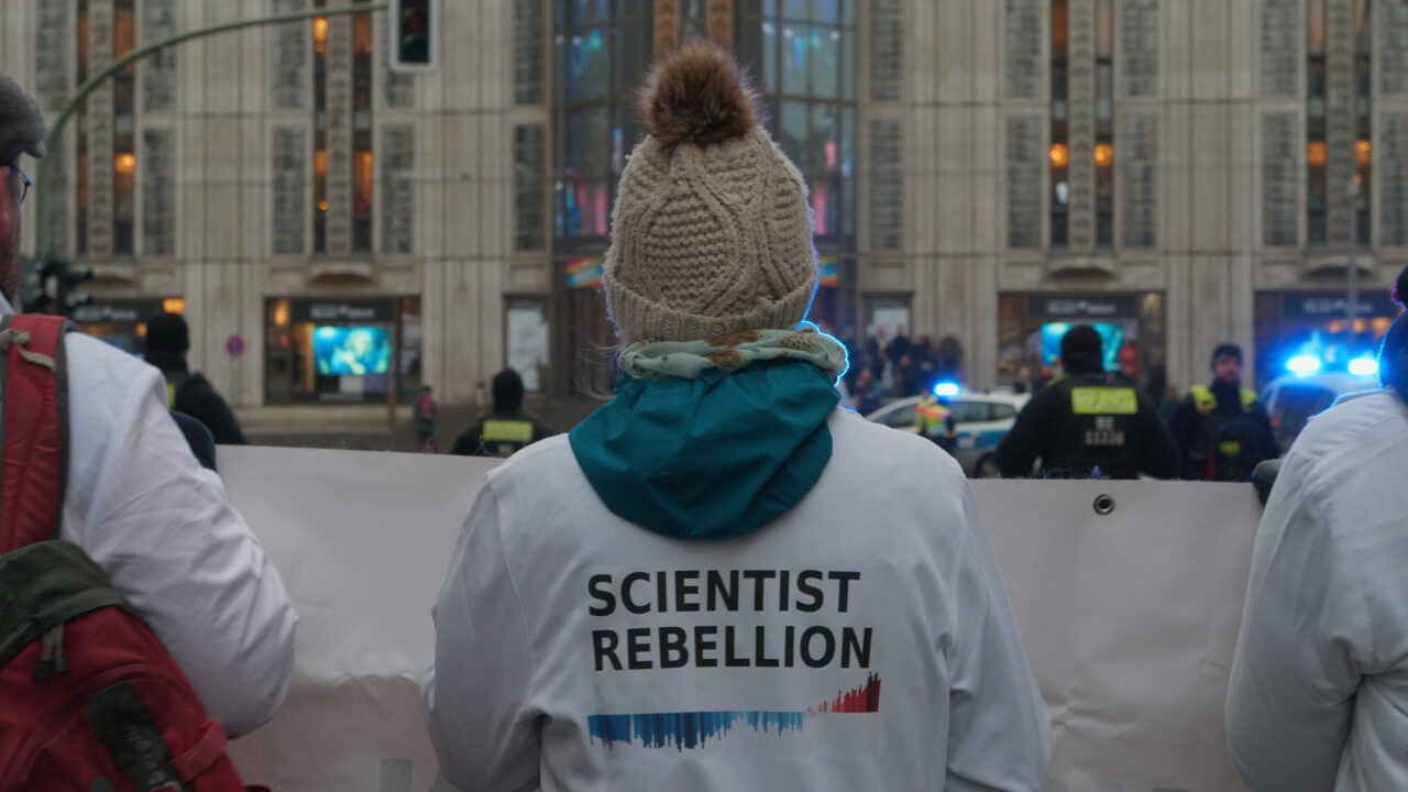Auf dem Foto ist eine Demonstration im öffentlichen Raum zu sehen. Im Mittelpunkt steht eine Person mit dem Rücken zur Kamera. Sie trägt eine weiße Jacke mit der Aufschrift „Scientist Rebellion“ sowie eine gestrickte Mütze mit Bommel und einen türkisfarbenen Kapuzenpullover. Die Person hält zusammen mit anderen Demonstrierenden ein großes weißes Transparent. Im Hintergrund befindet sich eine städtische Gebäudefassade mit großen Fenstern. Mehrere Polizeikräfte stehen gegenüber der Gruppe, teilweise sind blaue Lichter von Einsatzfahrzeugen zu erkennen. Die Szene wirkt wie eine öffentliche Protestaktion in einer Innenstadt.