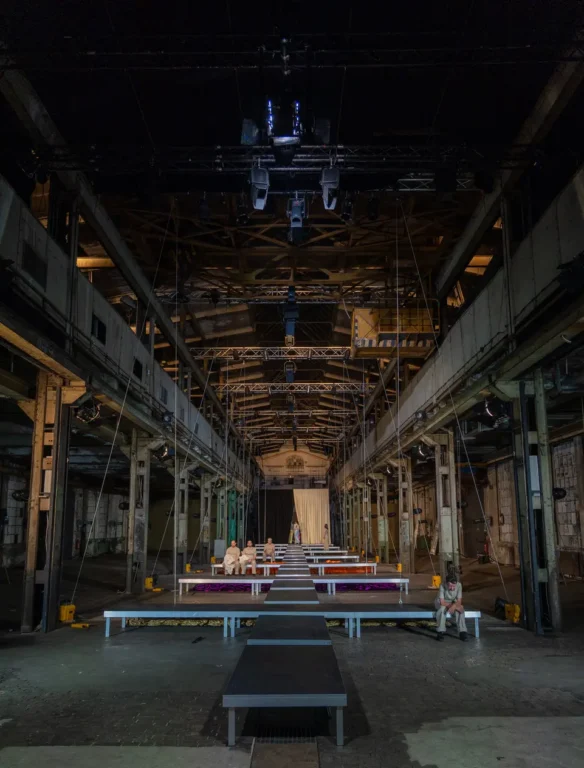 Eine weitläufige, historische Industriehalle mit freiliegender Stahlkonstruktion (Naxoshalle). In der Mitte führt ein langer schwarzer Laufsteg durch den Raum zu einer Bühne; darüber hängen Scheinwerfer und Lautsprecher. Mehrere Personen sitzen entlang des Laufstegs. Es handelt sich um ein Stück des Theater Willy Praml in der Naxoshalle.