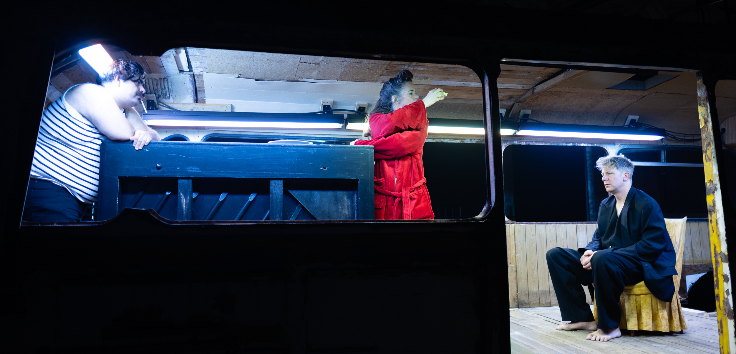 Three performers inside an old rusty bus: a man in a striped undershirt leans against a black piano, a woman in a red bathrobe stands beside him, and a barefoot man in a black suit sits at the right edge.