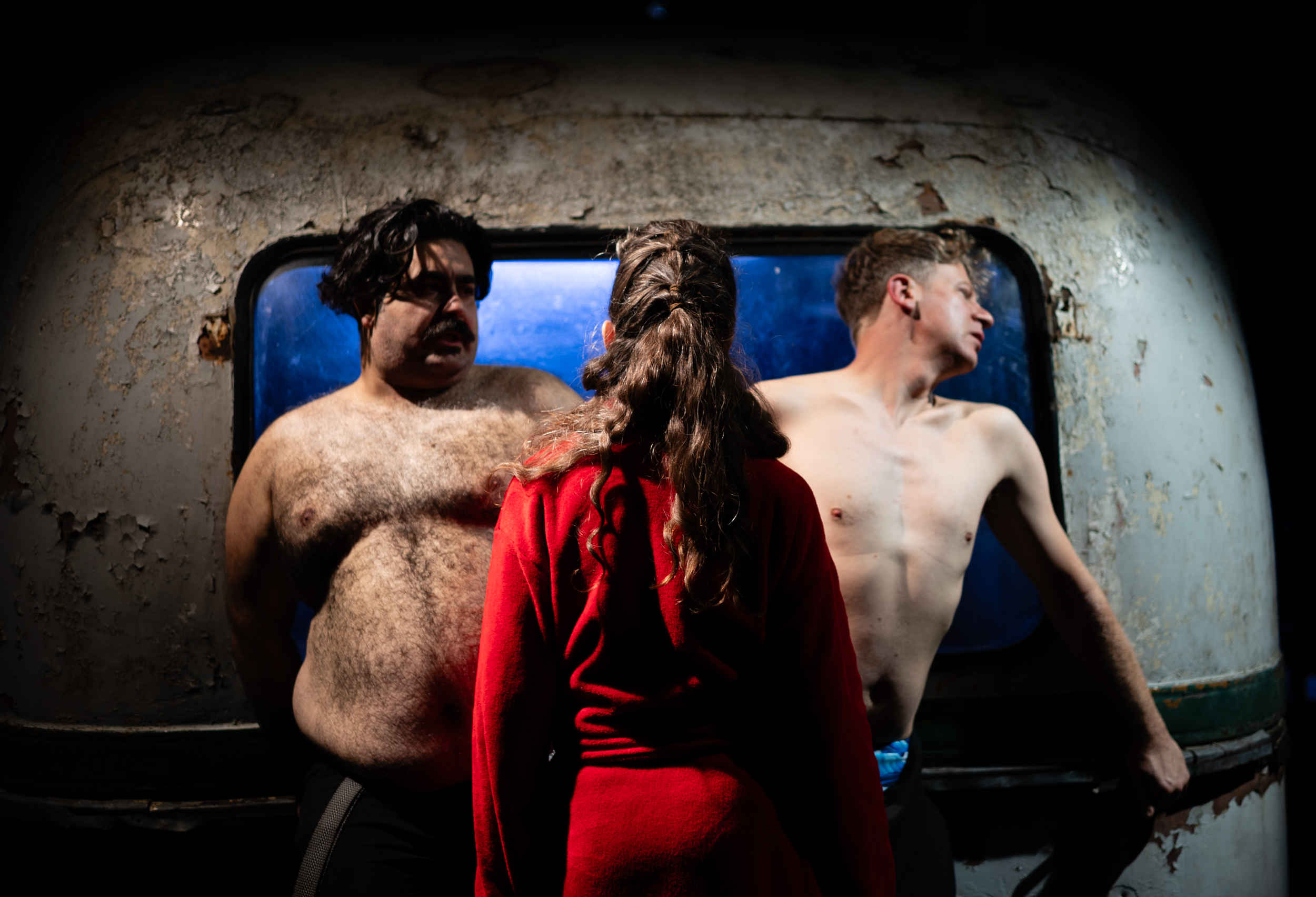 A woman in a red bathrobe stands between two shirtless men leaning against the wall of an old bus.