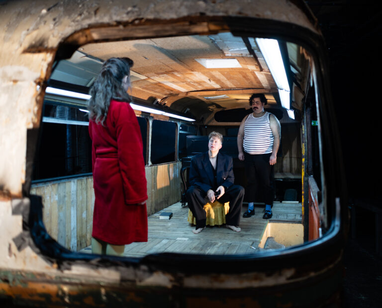 Drei Darsteller:innen in einem alten, verrosteten Bus: Eine Frau im roten Bademantel steht, ein barfüßiger Mann in schwarzem Anzug sitzt, ein Mann im gestreiften Unterhemd steht daneben.
