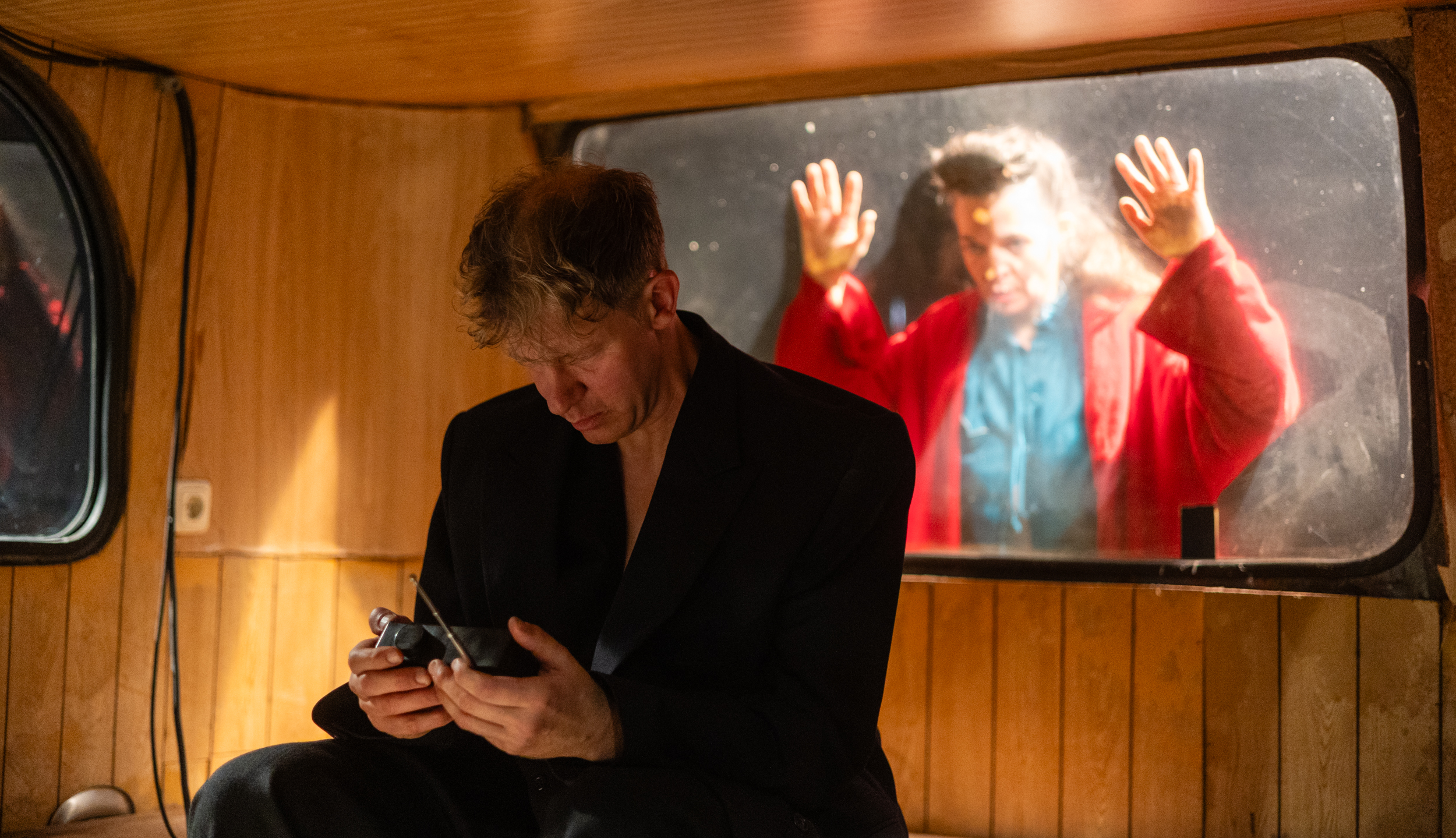 A man in a black suit sits pensively inside a wood-paneled bus, while outside, a woman in a red bathrobe presses her hands against the window.