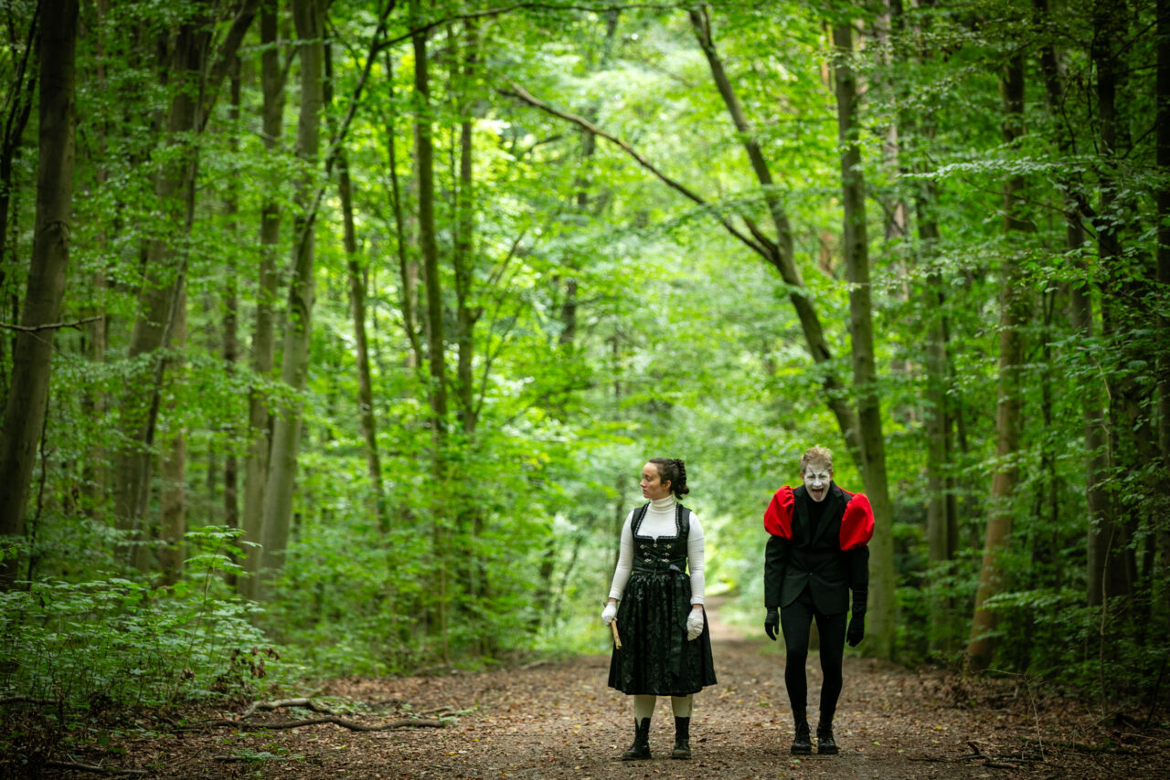 Two people in striking costumes stand on a forest path, surrounded by dense green foliage.