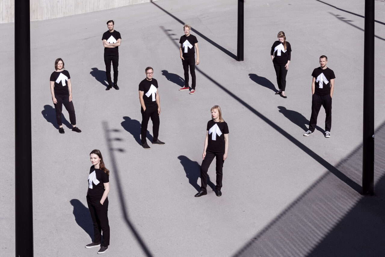 Acht Personen stehen mit Abstand zueinander im Freien, tragen schwarze T-Shirts mit weißem Pfeilmotiv.