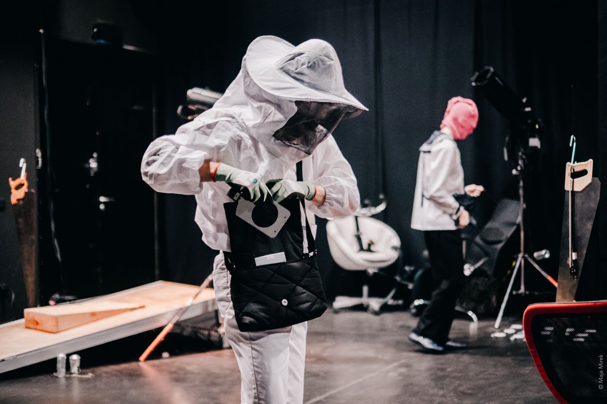 Two people in white protective suits and veiled hoods stand in a stage-like space, one working intently on a device.