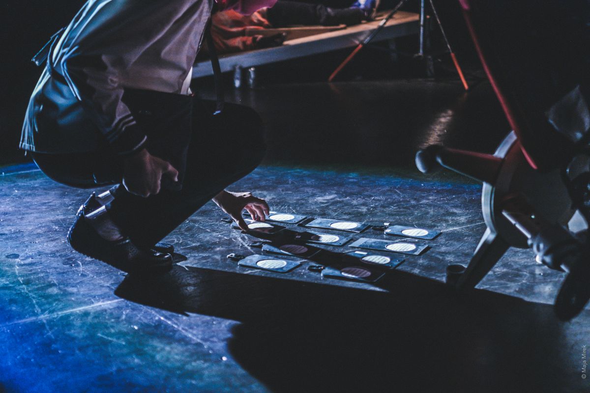 A person wearing a light jacket and dark trousers kneels on a stage, arranging flat circular objects on the floor while strong side lighting casts shadows.