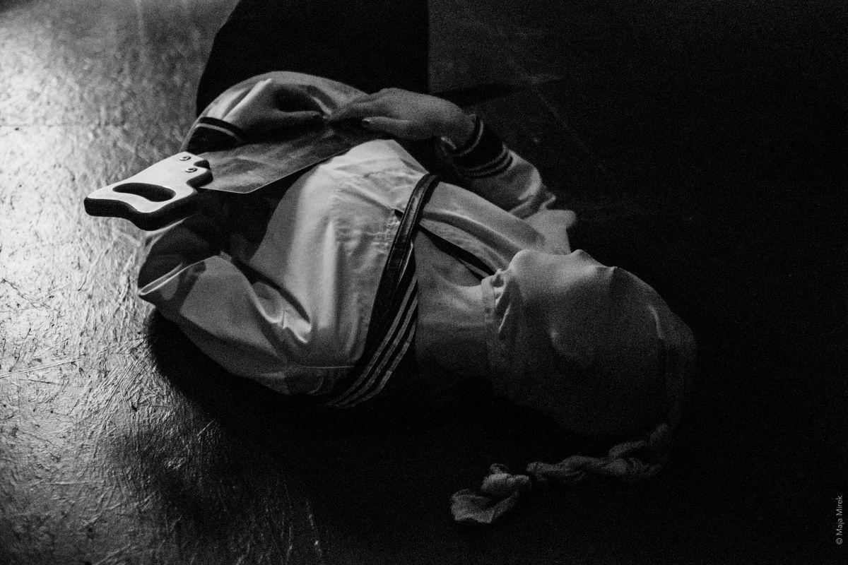 A person in a white jacket and hood lies motionless on the floor, their face covered by a mask. They loosely hold a flat metal blade.