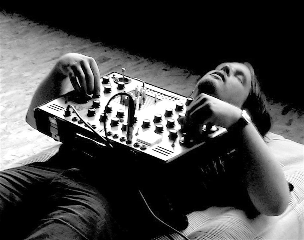 A person lies on their back on the floor and holds an audio mixing console on their chest. With both hands they adjust the knobs and controls. The photo is in black and white and has a calm, artistic atmosphere.