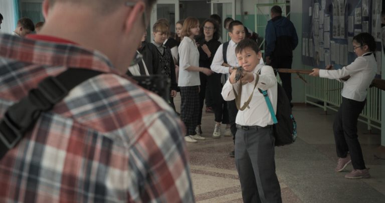 Ein Schulflur voller Kinder während einer Pause. Im Vordergrund steht unscharf eine Person mit kariertem Hemd und Rucksack. In der Mitte zielt ein Junge mit einem langen Spielzeuggewehr direkt nach vorne und kneift dabei ein Auge zu. Rechts daneben hält ein weiteres Kind ebenfalls ein langes Spielzeuggewehr. Im Hintergrund stehen mehrere Schülerinnen und Schüler in Gruppen zusammen und unterhalten sich. Die Kinder tragen überwiegend helle Oberteile und Schulkleidung.