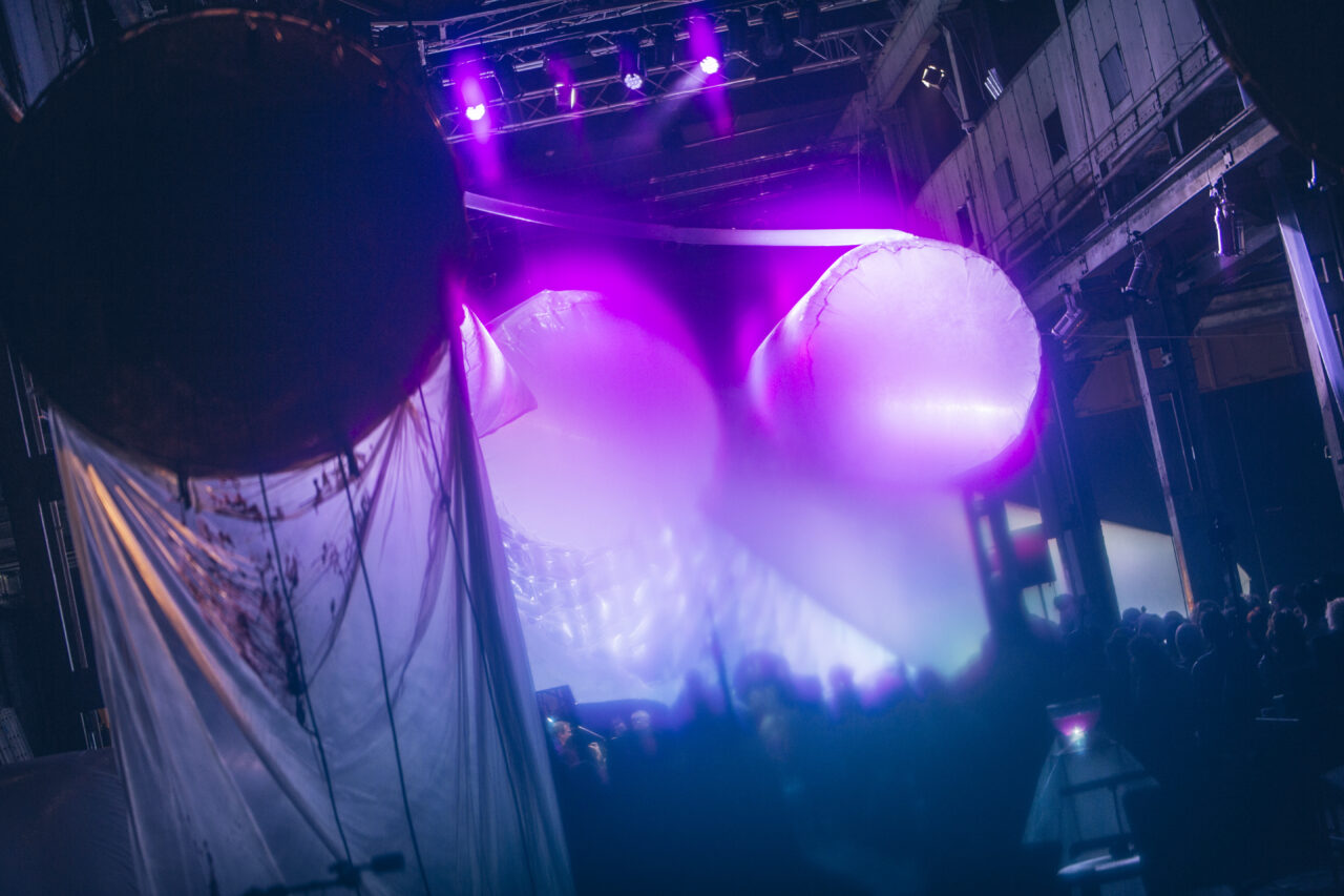 Large, glowing inflatable shapes hang on a stage under purple light, with an audience watching in the dim background.