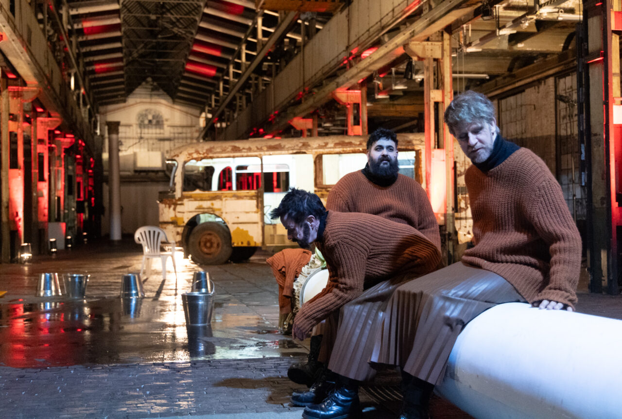 Drei Männer in orangebraunen Strickpullovern und langen Röcken sitzen in einer Industriehalle auf einem weißen Zylinder. Hinter ihnen steht ein alter, verrosteter Bus.