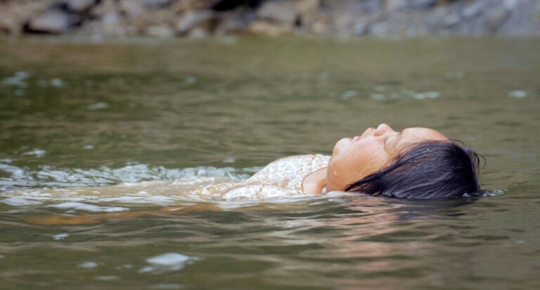 Eine Person treibt ruhig auf dem Rücken in einem flachen Gewässer. Der Kopf liegt leicht nach hinten geneigt im Wasser, die Augen sind geschlossen. Dunkles Haar breitet sich teilweise auf der Wasseroberfläche aus. Die Person trägt helle Kleidung mit dezentem Muster. Sanfte Wellen umgeben den Körper, während das unscharfe Ufer im Hintergrund in natürlichen Grau- und Grüntönen erscheint. Die Szene wirkt still, schwerelos und nach innen gekehrt und vermittelt eine Atmosphäre von Ruhe, Loslassen und Verletzlichkeit.