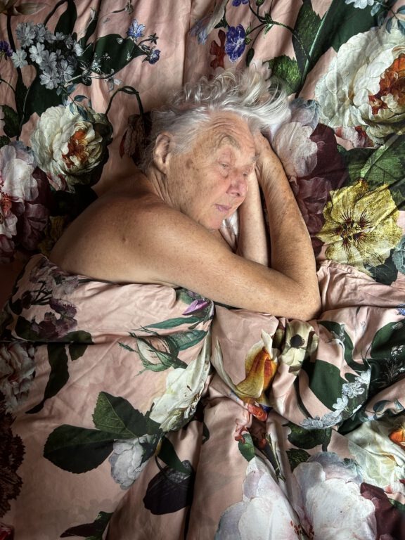 Eine ältere Person mit langen weißen Haaren liegt seitlich in einem Bett unter einer großflächig geblümten Bettdecke in Rosa-, Grün-, Weiß- und Violetttönen. Der nackte Oberkörper ist bis zu den Schultern sichtbar und teilweise unbedeckt, die Arme sind unter dem Kopf angewinkelt. Die Person wirkt ruhig und entspannt. Bettwäsche und Hintergrund gehen durch das durchgehende florale Muster nahezu ineinander über.
