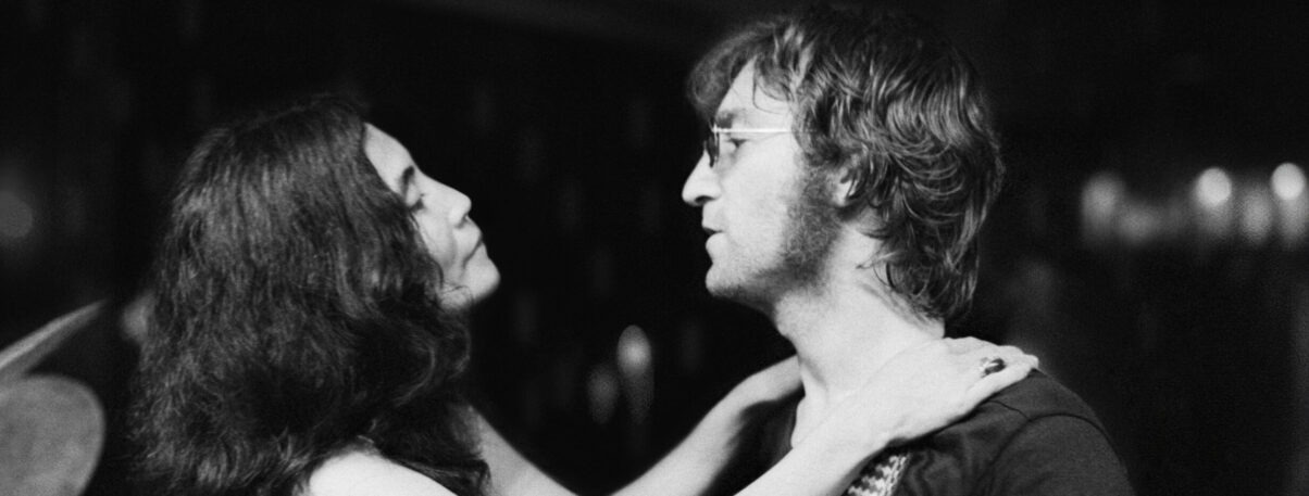 Das Schwarz-Weiß-Bild zeigt Yoko Ono und John Lennon in einer Nahaufnahme. Yoko Ono legt beide Hände auf John Lennons Schultern, während sich die beiden aufmerksam ansehen. John Lennon trägt eine Brille und hat mittellanges, leicht welliges Haar. Die Szene wirkt ruhig und intim. Der dunkle, unscharfe Hintergrund lenkt den Blick auf die beiden Personen und ihren gemeinsamen Moment.