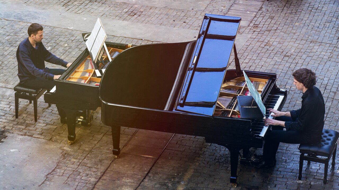Two pianists sit facing each other at two grand pianos, playing. Both wear black clothing, with sheet music on the stands.