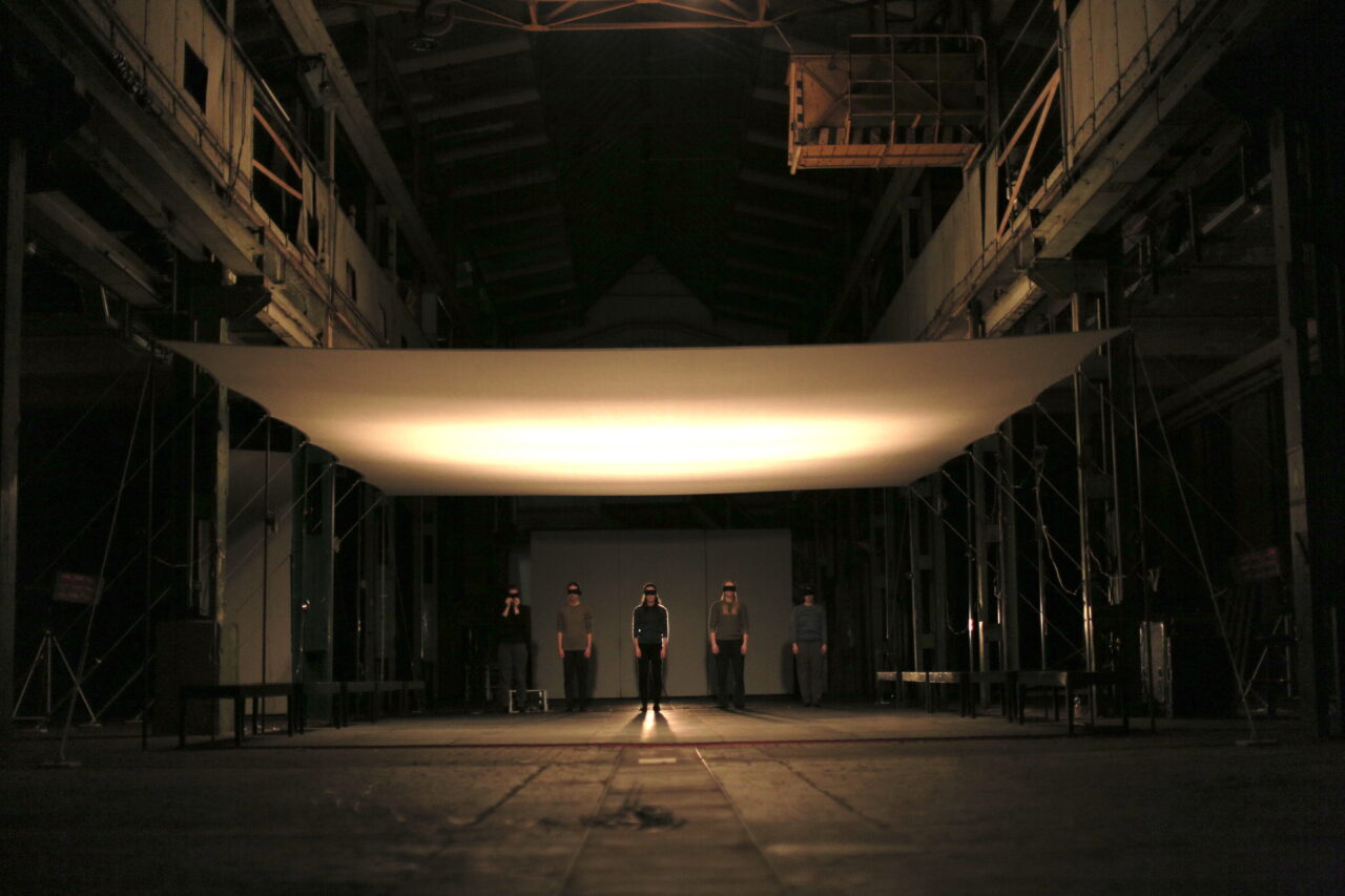 Fünf Personen stehen in einer Reihe auf einer Bühne in einer großen, dunklen Industriehalle. Über ihnen hängt eine breite, helle Stoffbahn, die von Scheinwerfern angestrahlt wird.