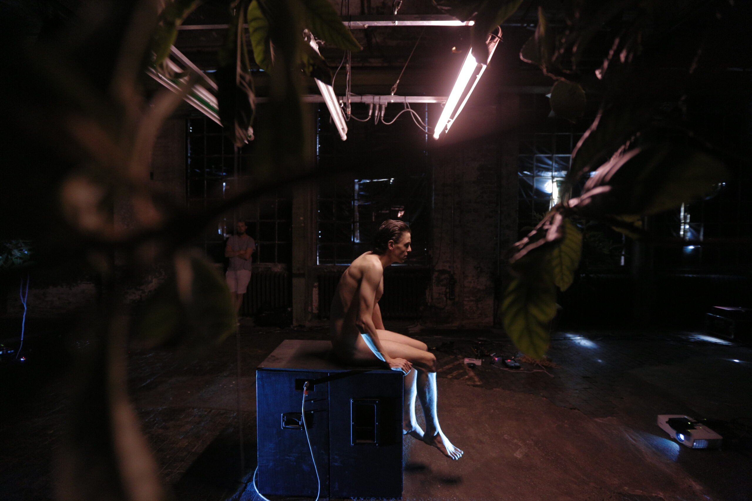 Ein nackter Mann sitzt auf einem schwarzen Kasten in einer dunklen Industriehalle, nur von Neonröhren über ihm beleuchtet.