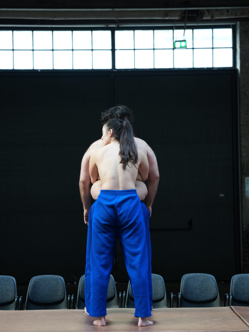 Zwei Personen mit nacktem Oberkörper stehen eng umschlungen auf einer Bühne. Beide tragen blaue Hosen und stehen barfuß, mit dem Rücken zum Publikum.