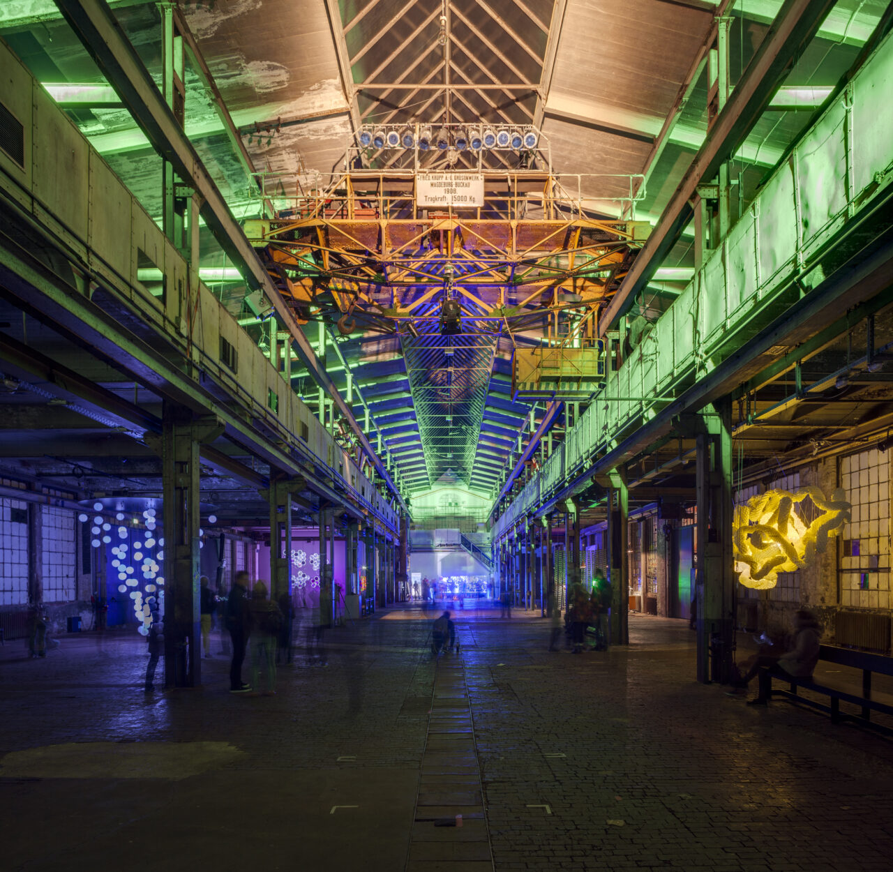 Eine Gruppe von Menschen in auffälligen Kostümen steht an einer Bushaltestelle im Wald vor einem großen Fahrplan.