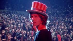 Das Bild zeigt eine Konzertszene aus der Perspektive der Bühne. Im Vordergrund steht eine Person (Mick Jagger) mit einem rot-weiß gestreiften Zylinderhut und einem Mikrofon vor dem Mund. Die Person ist seitlich zu sehen und wird von rötlich-blauem Bühnenlicht beleuchtet. Im Hintergrund befindet sich eine große, dicht gedrängte Menschenmenge, die zur Bühne blickt. Die Atmosphäre wirkt lebhaft und musikalisch, vermutlich während eines Live-Auftritts.