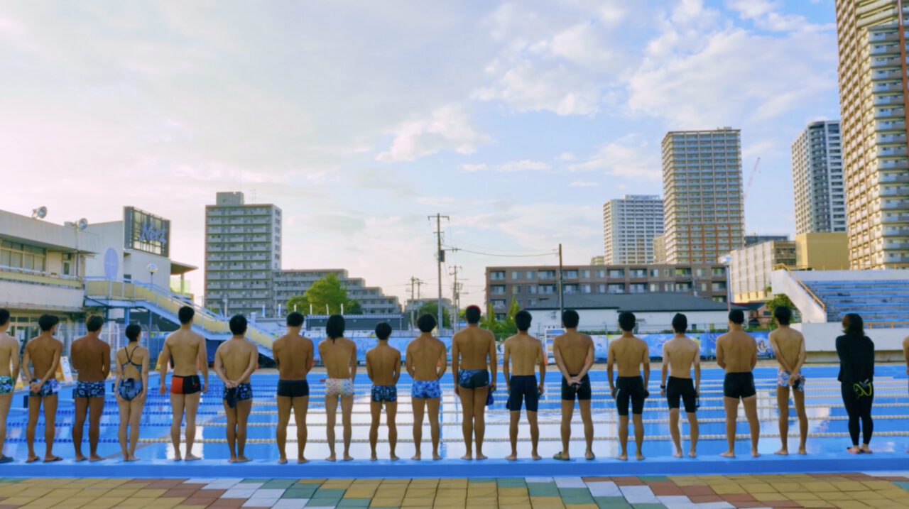 Eine Gruppe von Schwimmerinnen und Schwimmern steht nebeneinander am Rand eines großen Außenpools und blickt auf das Wasser. Im Hintergrund erheben sich moderne Hochhäuser unter einem hellen Himmel mit leichter Abendstimmung. Die Szene vermittelt Teamgeist, Vorbereitung und urbanes Sportleben.