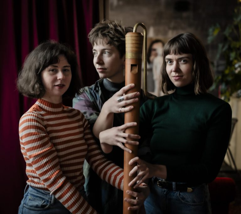Three women stand close together, holding a large wooden flute upright, looking calmly at the camera.